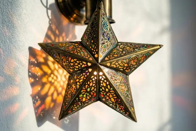 Een stervormige messing lantaarn met fijne opengewerkte patronen werpt kleurrijke schaduwen op een witte muur in warm goudkleurig licht.