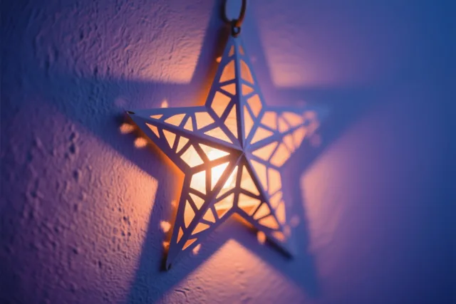 Een hangende sterlantaarn met een geometrisch uitgesneden patroon werpt warm licht op een muur met structuur in een blauwpaarse omgeving.