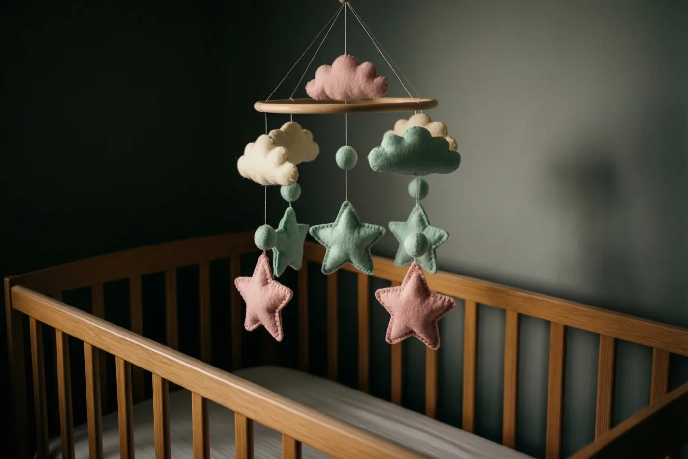 Een houten mobiel met vilten sterren en wolken hangt boven een leeg houten babybedje tegen een donkere muur in zacht licht.