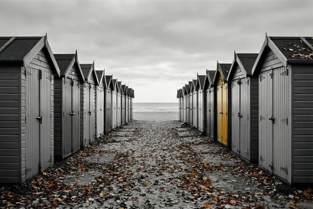 Gesloten strandhuisjes staan symmetrisch aan weerszijden van een zandpad naar de zee onder een grijze hemel, met één felgele deur tussen grijze en bruine tinten.