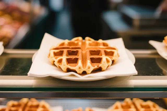 Een goudbruine suikerwafel ligt op een wit servet op een klein bord op een glazen toonbank in warm licht.