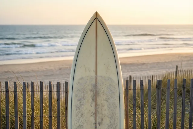 Een surfplank staat rechtop tegen een houten duinhek, met een zandstrand en zachte golven erachter in het gouden licht van de zonsopgang.