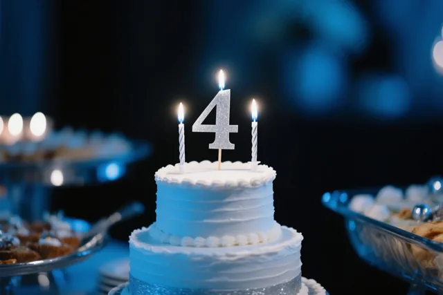 Een taart met meerdere verdiepingen, wit glazuur en blauwe accenten staat op een buffet met een glitterend cijfer 4 en drie brandende kaarsjes onder blauw licht.