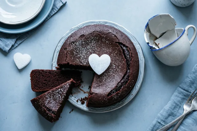 Een chocoladetaart met een suikerhart waaruit twee stukken ontbreken, staat op een bord naast een gebroken wit kopje met blauwe rand op een blauw tafelkleed.
