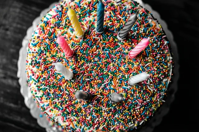 Van bovenaf zie je een ronde taart vol regenboogkleurige suikerstrooisels en verschillende kaarsjes op een wit bord met geschulpte rand tegen een donkere achtergrond.