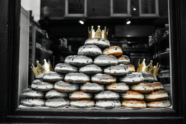 Een piramide van driekoningentaarten met bloemsuiker en gouden papieren kronen staat in een bakkersetalage, met de taarten en kronen in kleur tegen een zwart-witte achtergrond.
