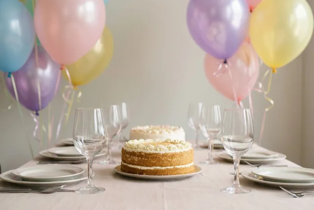 Een gedekte tafel met een gelaagde taart, wijnglazen en borden is versierd met pastelroze, blauwe, gele en paarse ballonnen.