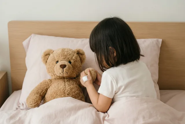 Een meisje met donker haar zit met de rug naar de kijker in bed en stopt een bruine teddybeer onder roze dekens, bij een licht houten hoofdbord.