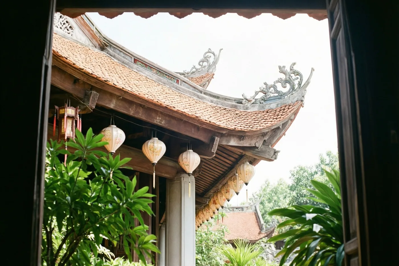 Van onderaf zijn gebogen dakranden met pannen en hangende papieren lantaarns aan een tempel te zien, omlijst door donkere houten deuropeningen en groen tegen een heldere hemel.