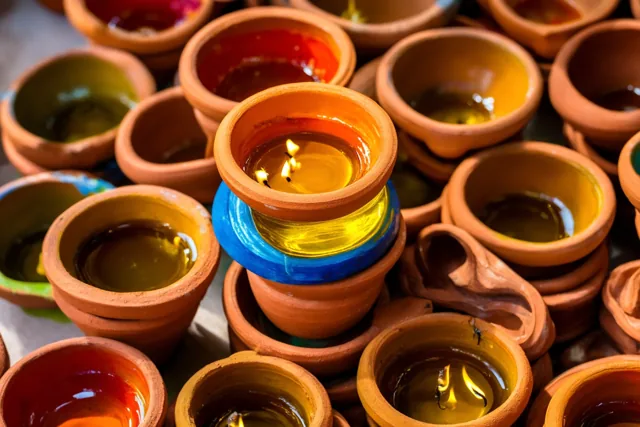 Op een stapel terracotta olielampjes brandt één kleine vlam, omringd door onverlichte lampjes in verschillende kleuren met gele en blauwe accenten.
