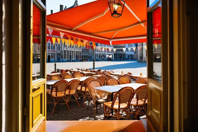 Vanuit een café is een zonnig terras op een plein te zien, met tafels en stoelen onder een groot oranje zonnescherm en kleurrijke vlaggetjes.