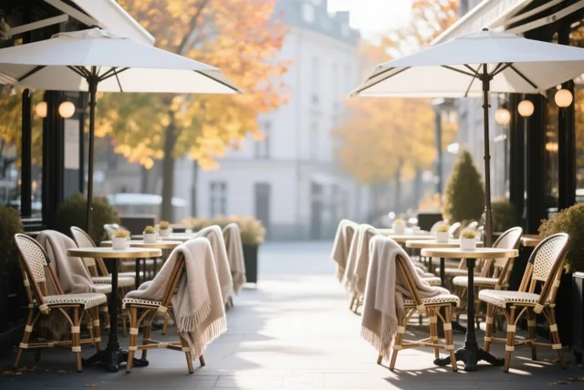 Stoelen met beige dekens staan op een caféterras voor bomen met gouden herfstbladeren in zacht warm licht.