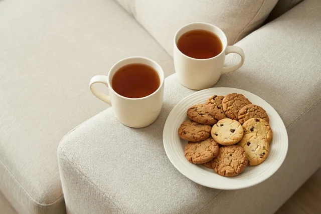 Twee witte mokken thee en een bord koekjes met stukjes chocolade staan op de armleuning van een lichtbeige zetel.