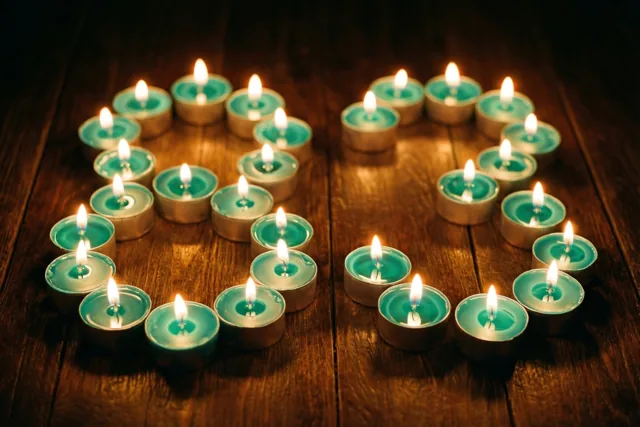 Brandende theelichtjes vormen 83 op een donkere houten vloer, in een beeld met blauwgroene, gouden en donkerbruine tinten.