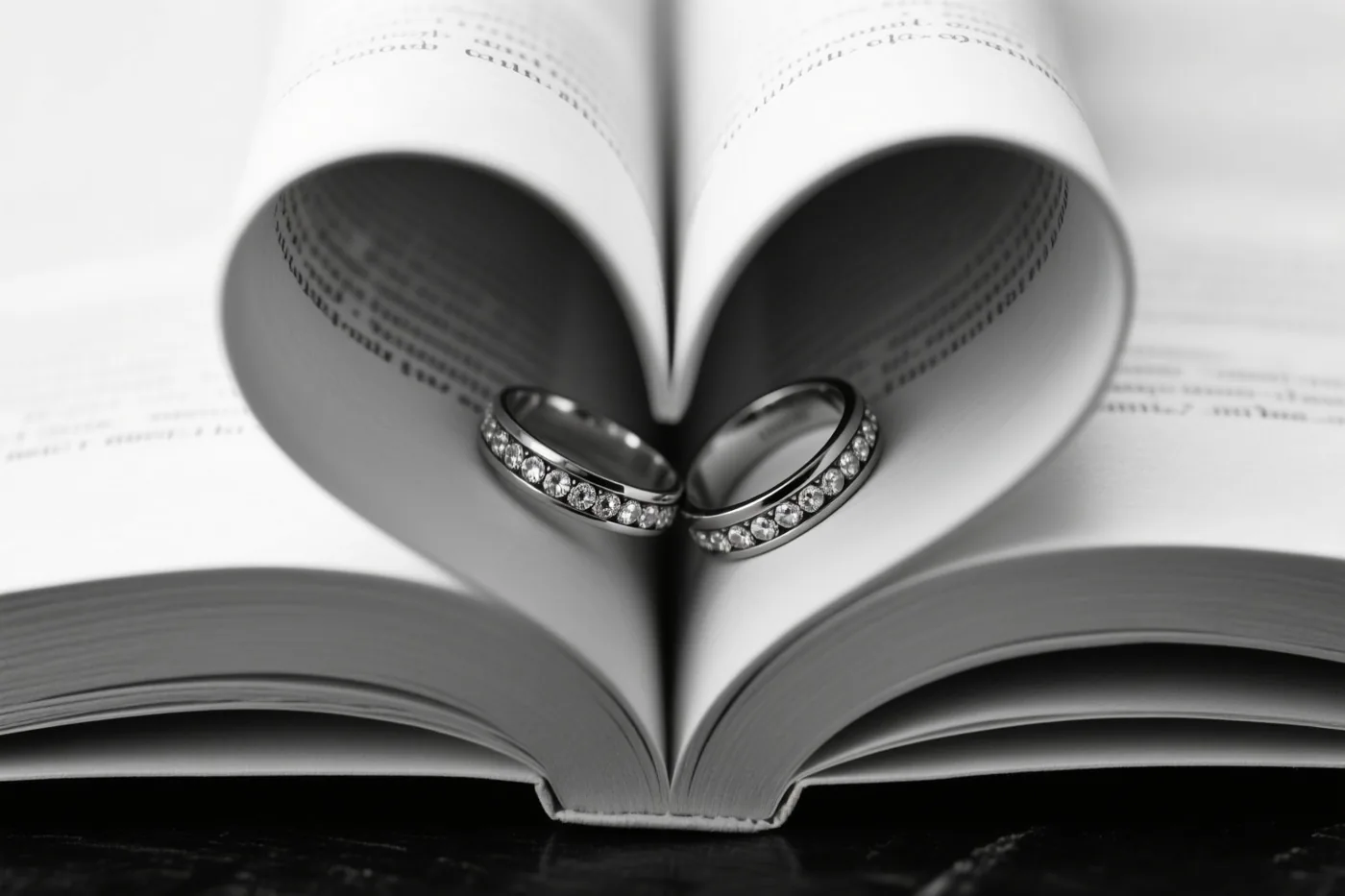 Een zwart-witfoto toont twee trouwringen met diamanten in een open boek waarvan de gebogen bladzijden een hart vormen.