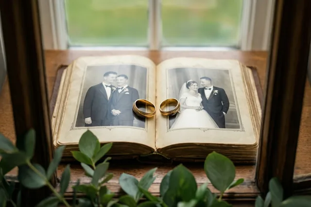 Twee gouden trouwringen liggen op een open oud fotoalbum met zwart-witte huwelijksportretten, op een houten ondergrond bij een raam met groen op de voorgrond.