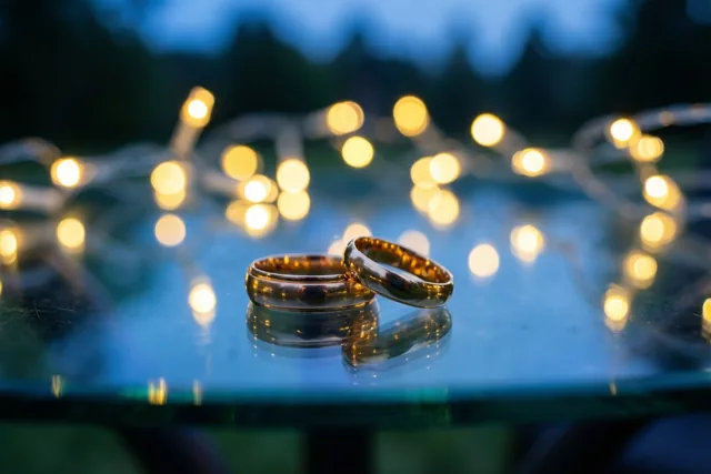 Twee gouden trouwringen liggen op een spiegelende glazen tafel voor onscherpe lichtjes, met een donkerblauwe achtergrond en warme gouden accenten.