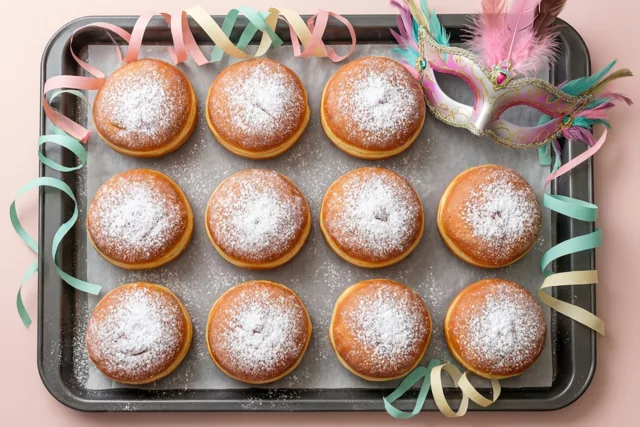 Twaalf ronde donuts met bloemsuiker liggen op een bakplaat tussen gekleurde papieren slingers en een verenmasker, tegen een zachtroze achtergrond.