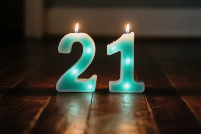 Twee brandende theelichtkaarsen in de vorm van de cijfers 2 en 1 staan op een donkere houten vloer, in blauwgroen en wit licht.