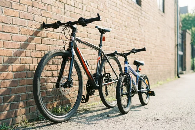 Een grote fiets en een kleine kinderfiets leunen op een verharde ondergrond tegen een bakstenen muur in warm zonlicht.