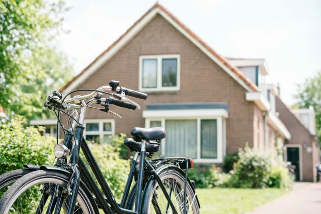 Twee fietsen staan naast elkaar voor een bakstenen rijwoning met een trapgevel in een zonnige, groene woonbuurt.