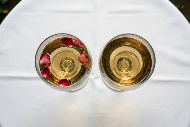 Van bovenaf zijn twee champagneglazen met goudkleurige drank en rozenblaadjes op een wit tafelkleed te zien.