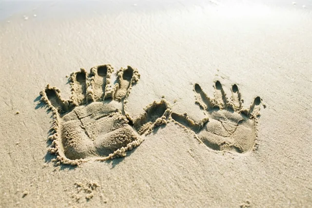 Een grote en een kleine handafdruk staan naast elkaar in nat zand, beschenen door warm zonlicht.