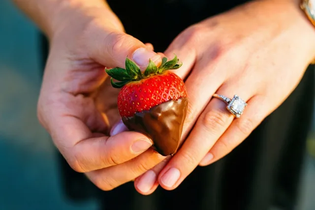 Twee handen houden voorzichtig een in chocolade gedoopte aardbei vast, met een opvallende verlovingsring aan de ringvinger in warm licht.