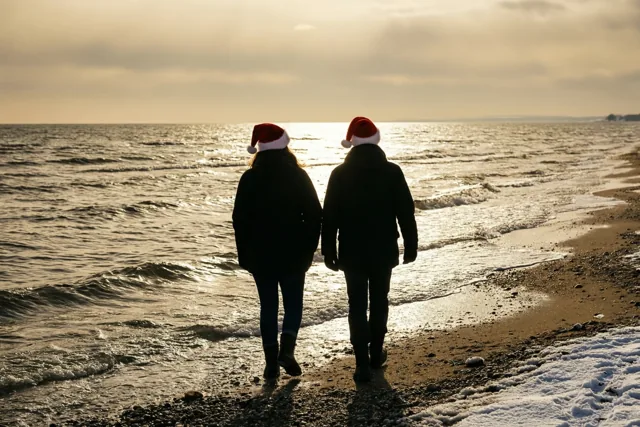 Twee mensen met kerstmutsen wandelen met hun rug naar de camera over een besneeuwd strand langs een winterse zee in warm gouden licht.