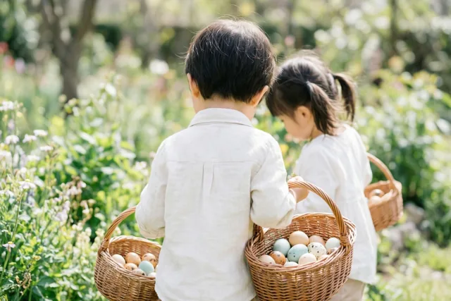 Twee kinderen lopen met hun rug naar de camera door een zonnige tuin, elk met een rieten mand gekleurde eieren, tussen groen en witte bloemen.