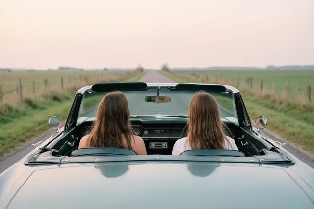 Twee vrouwen met lang haar zitten in een oude groene cabrio op een rechte landweg, van achteren gezien in zacht, warm licht.
