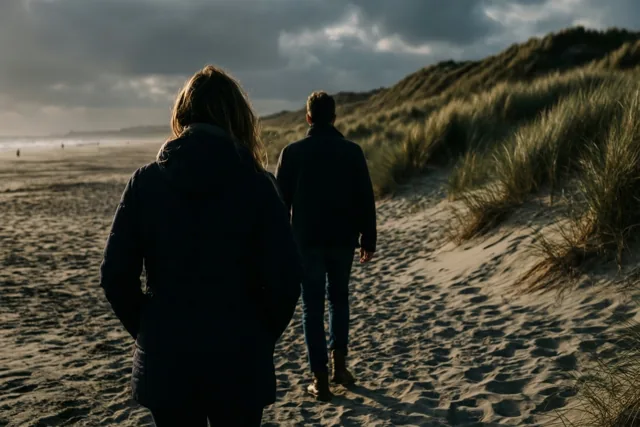 Twee mensen lopen met hun rug naar de kijker over een zandstrand naar met gras begroeide duinen onder een bewolkte hemel.