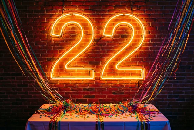 Een groot neongetal 22 straalt aan een rode bakstenen muur boven een tafel met kleurrijke slingers en confetti.
