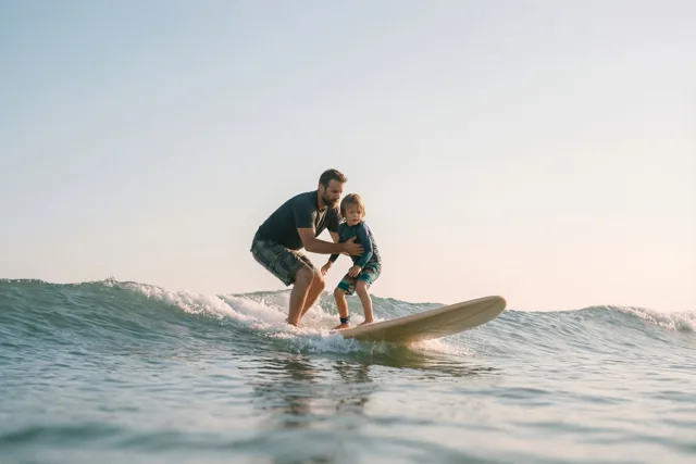 Een vader ondersteunt zijn jonge kind op een surfplank terwijl ze samen op een kleine zeegolf surfen onder een heldere hemel.