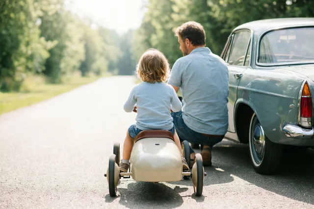 Een kind rijdt in een retrotrapauto naast zijn vader, die bij een echte oldtimer knielt, beiden van achteren gezien op een zonnige verharde landweg met bomen.