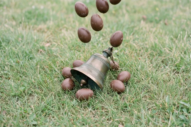Een klein bronzen klokje ligt in groen gras, omringd door chocolade-eieren die uit de lucht lijken te vallen.