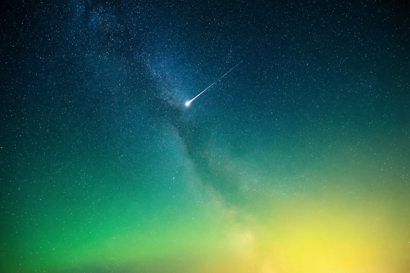 Een vallende ster trekt door een sterrenhemel met blauwgroene, groene en gele tinten bij de horizon.