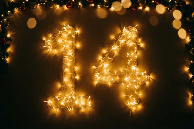 Warme lichtjes vormen 14 tegen een donkere muur, omlijst door een feestelijke slinger met onscherpe lichtjes op de achtergrond.