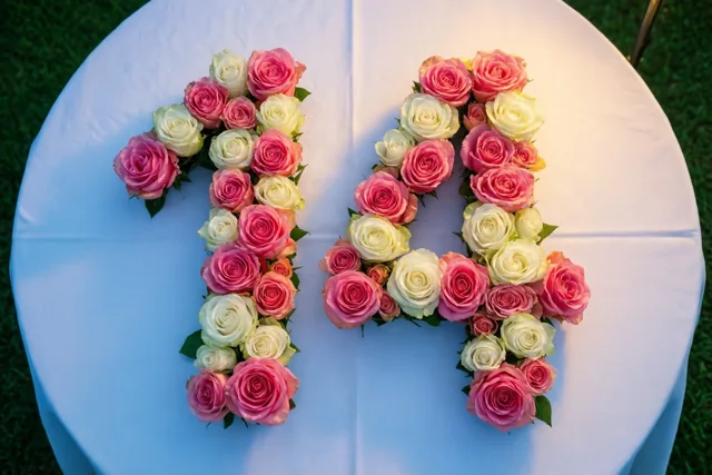 Roze en witte rozen vormen 14 op een wit tafelkleed buiten op groen gras, van bovenaf gezien.