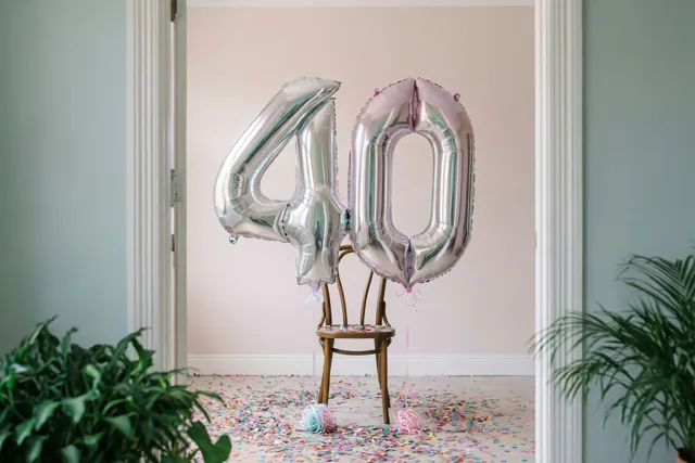 Twee grote metaalglanzende ballonnen vormen 40 aan een houten stoel in een helder verlichte kamer met kleurrijke confetti op de vloer en planten naast de deuropening.