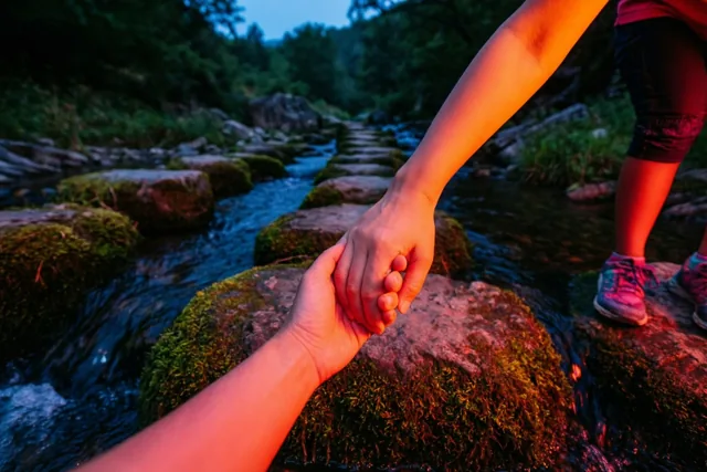Een grote en een kleine hand houden elkaar vast boven bemoste stenen in een bosbeek, met warm oranje licht tegen een koelblauwe achtergrond in de schemering.