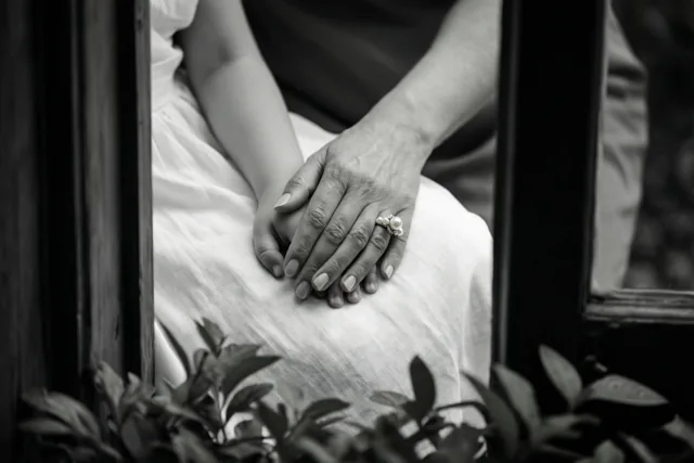 Een zwart-witdetailfoto toont een volwassen hand met een parelring die een kinderhand vasthoudt op wit linnen, omlijst door donkere houten spijlen en onscherp gebladerte op de voorgrond.