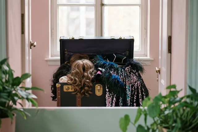Een open zwarte koffer vol kleurrijke pruiken, veren en capes met pailletten staat voor een raam in een kamer met roze muren en groene planten op de voorgrond.