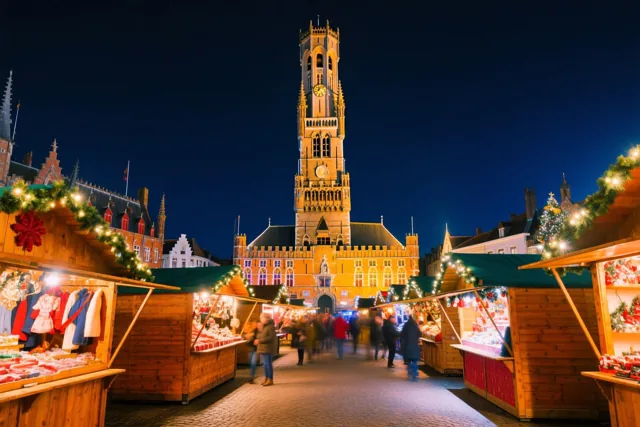 Houten kraampjes met lichtjes en slingers staan op een Brugse kerstmarkt onder een diepblauwe nachtelijke hemel, met het verlichte Belfort op de achtergrond.