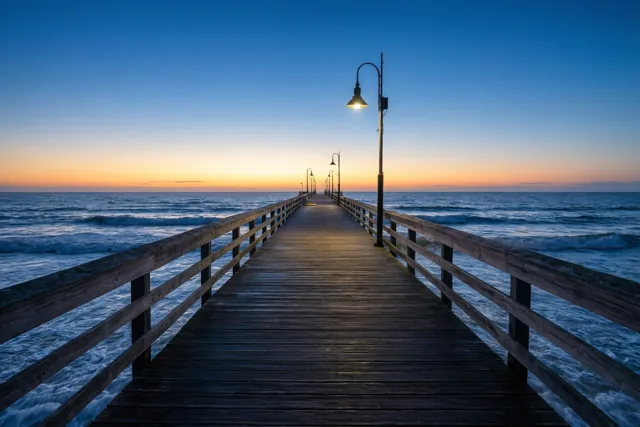Een lange houten pier met brandende lantaarns strekt zich uit over zacht golvend zeewater bij zonsopgang, onder een hemel die van oranje aan de horizon naar diepblauw overgaat.