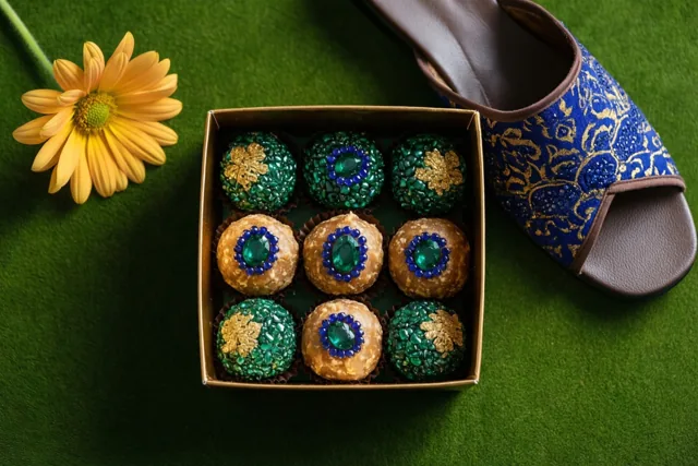 Een doos versierde pralines staat op een groene ondergrond naast een gele bloem en een blauw met gouden pantoffel.