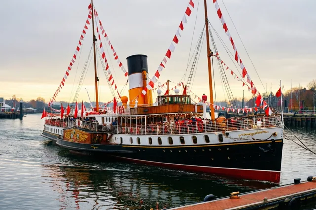 Een stoomboot met rode en witte vlaggen ligt op een grijze ochtend aan een havenkade, in zwarte, witte, rode en oranje tinten.