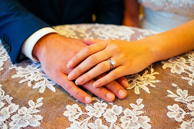 Een mannenhand en een vrouwenhand zijn verstrengeld boven een wit tafelkleed met bloemenkant, met een opvallende diamanten verlovingsring aan de vrouwenhand in zacht warm licht.