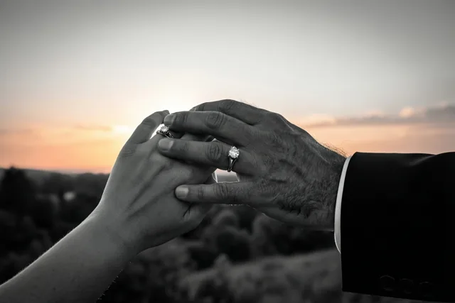 Twee handen grijpen in elkaar met een verlovingsring aan één vinger, in een zwart-witbeeld met een warme gloed van de zonsondergang erachter.