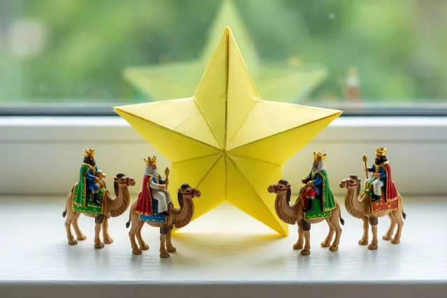 Vier miniatuurfiguurtjes van koningen op kamelen staan op een vensterbank naar een grote gele papieren ster gericht, in warm licht voor een wazige groene achtergrond.